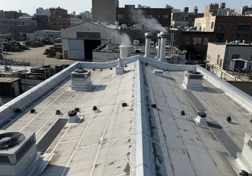 A commercial rooftop with an older PVC roof membrane showing visible wear, along with multiple vents, HVAC units, and exhaust stacks surrounded by industrial buildings.